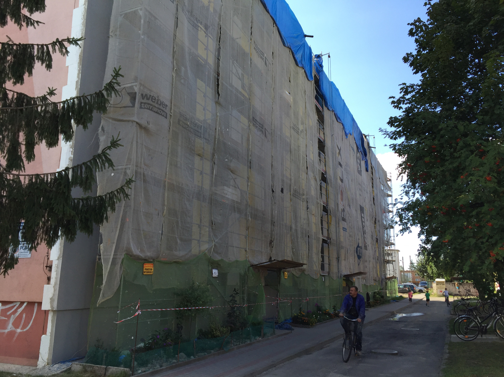 Wielopiętrowy budynek w trakcie ocieplania, osłonięty siatką z widocznym logo Weber Terranova, z niebieskim zabezpieczeniem na dachu, na pierwszym planie rowerzysta na ulicy.