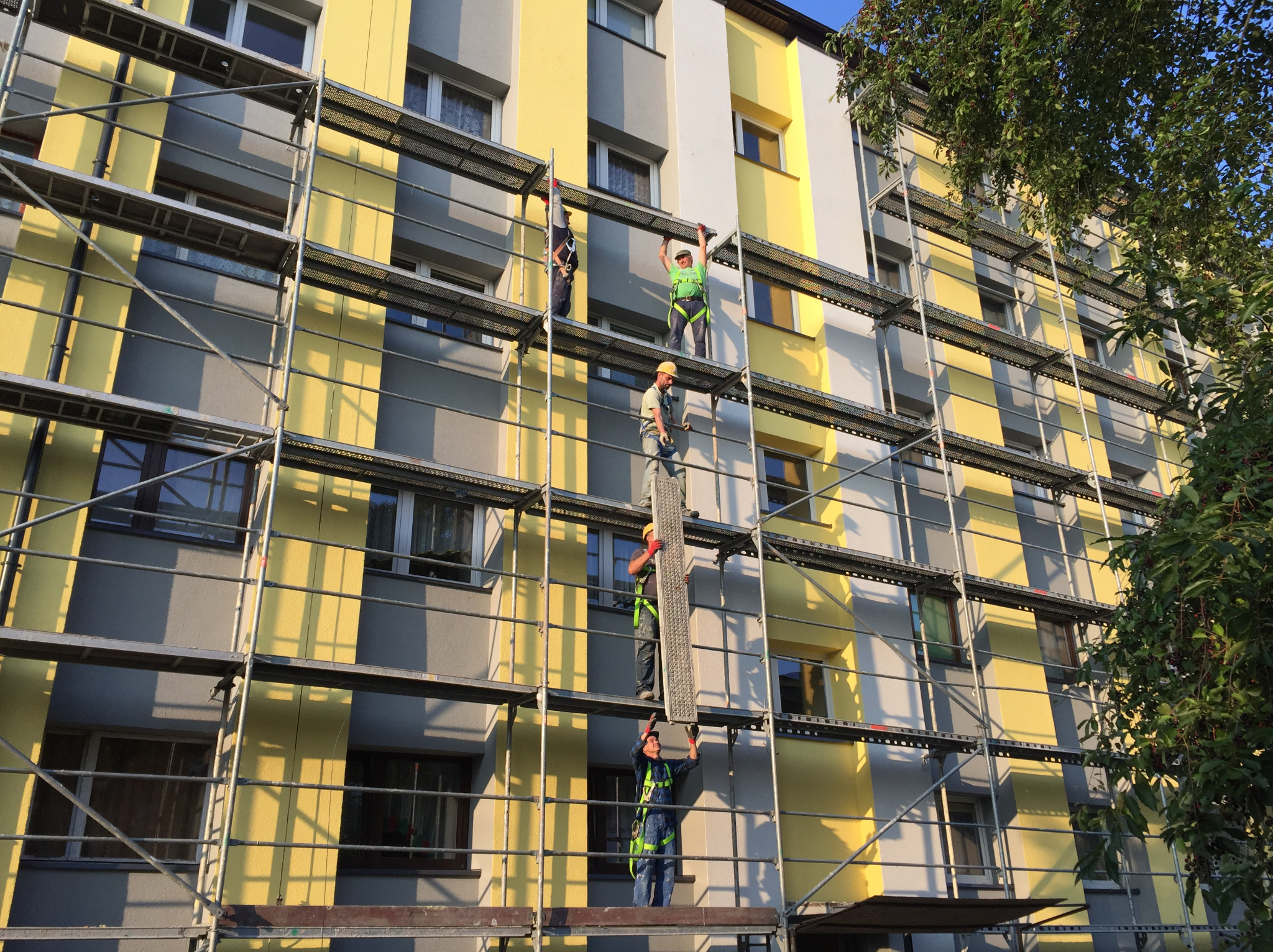 Pracownicy na rusztowaniu montują elementy elewacji budynku w odcieniach żółci i szarości.