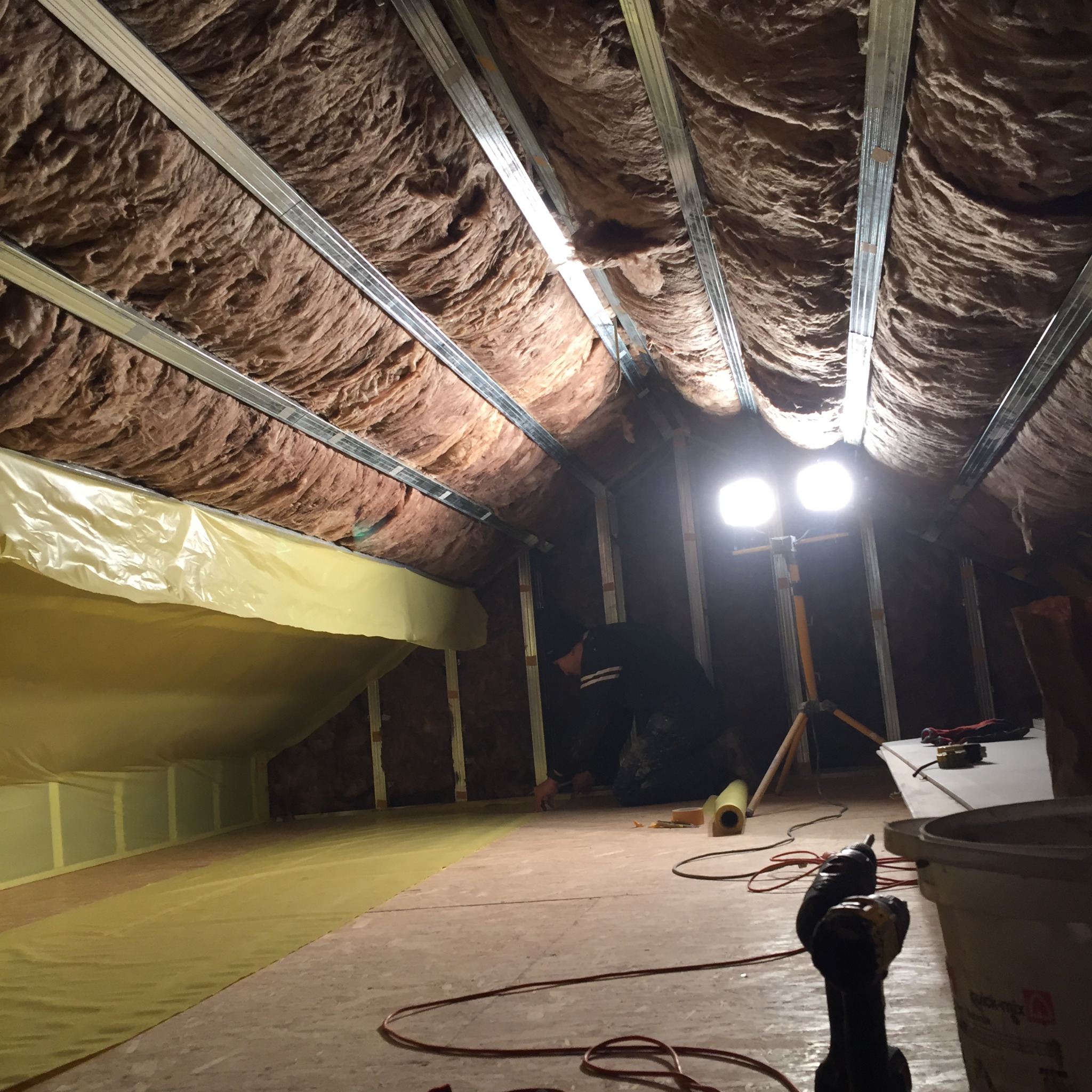 Poddasze w trakcie ocieplania wełną mineralną, widoczne metalowe profile, folia paroizolacyjna, narzędzia pracy i lampa oświetleniowa na statywie.