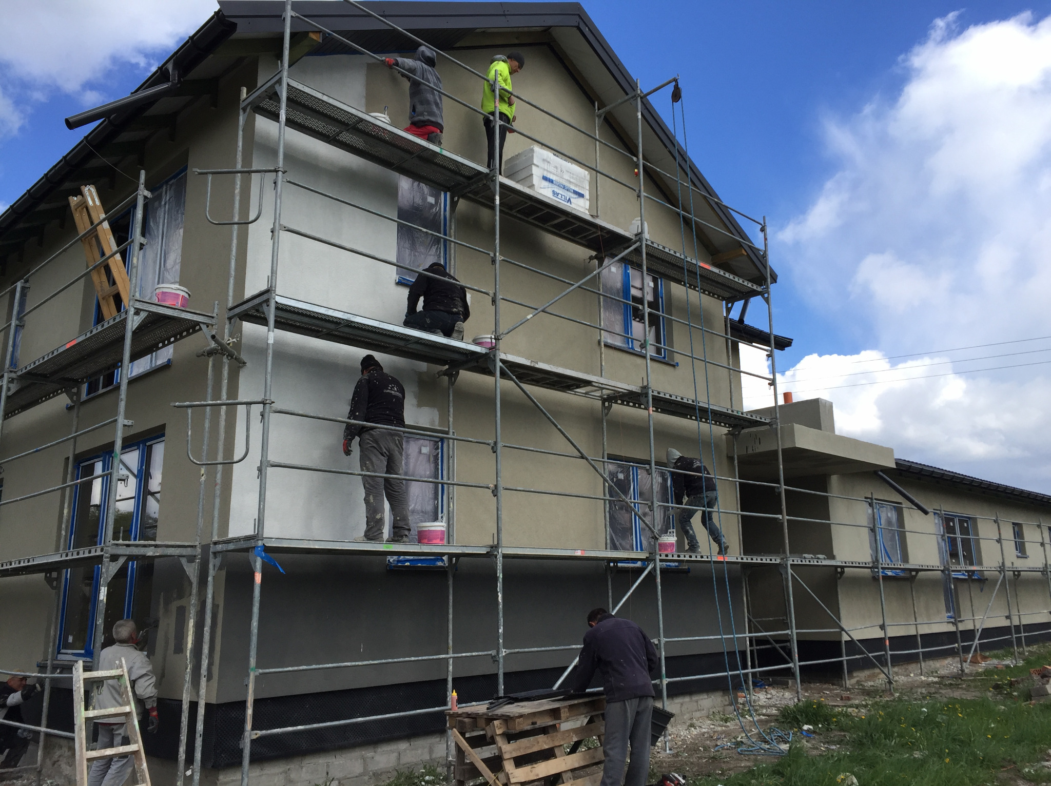Ekipa budowlana na rusztowaniu podczas nakładania tynku na elewację domu jednorodzinnego, okna zabezpieczone folią, widoczne puszki z farbą i paleta z materiałami.