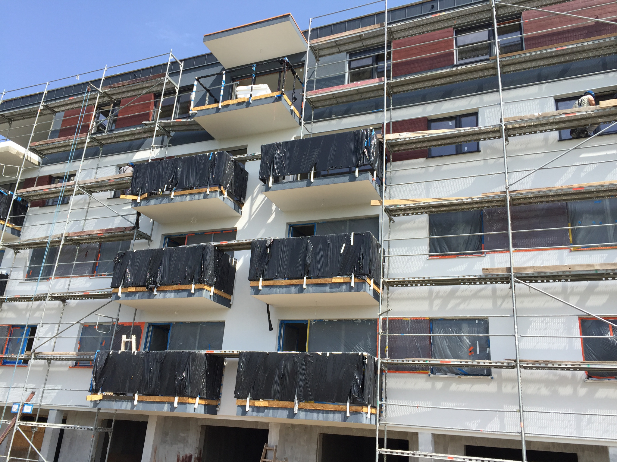 Wielopiętrowy budynek w trakcie prac elewacyjnych, zabezpieczone balkony folią, rusztowania na całej fasadzie, pracownik w oknie na górnym piętrze.