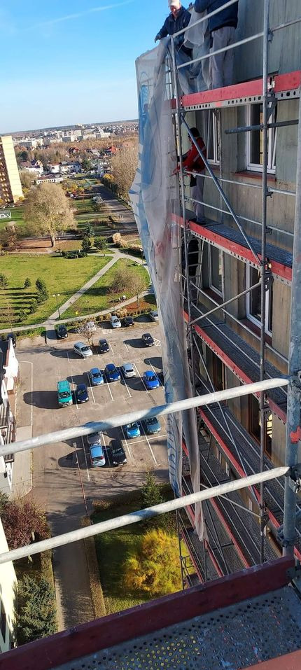 Prace na wysokościach: Robotnicy na rusztowaniu przy ocieplaniu bloku mieszkalnego, widok z góry na parking i park.