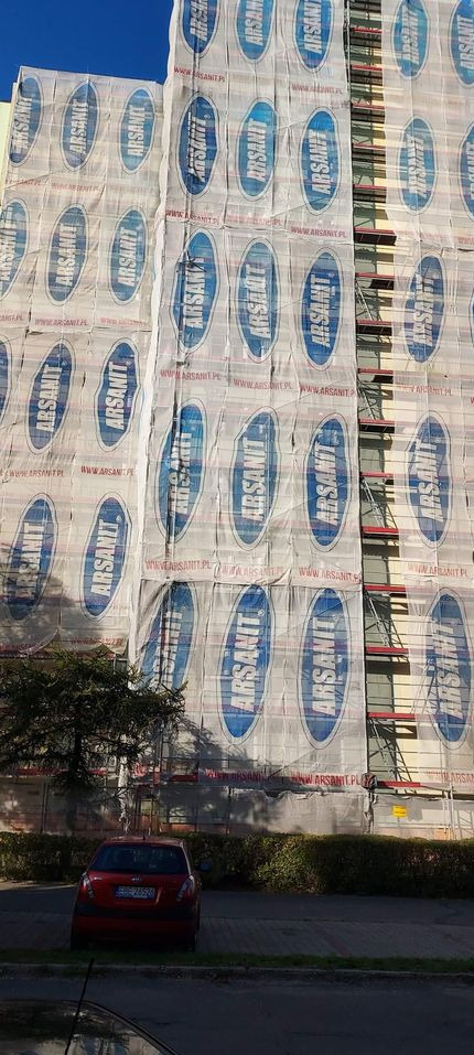 Wielopiętrowy budynek w trakcie ocieplania, pokryty rusztowaniami i siatką z logo Arsanit, widoczny fragment parkingu z czerwonym samochodem.