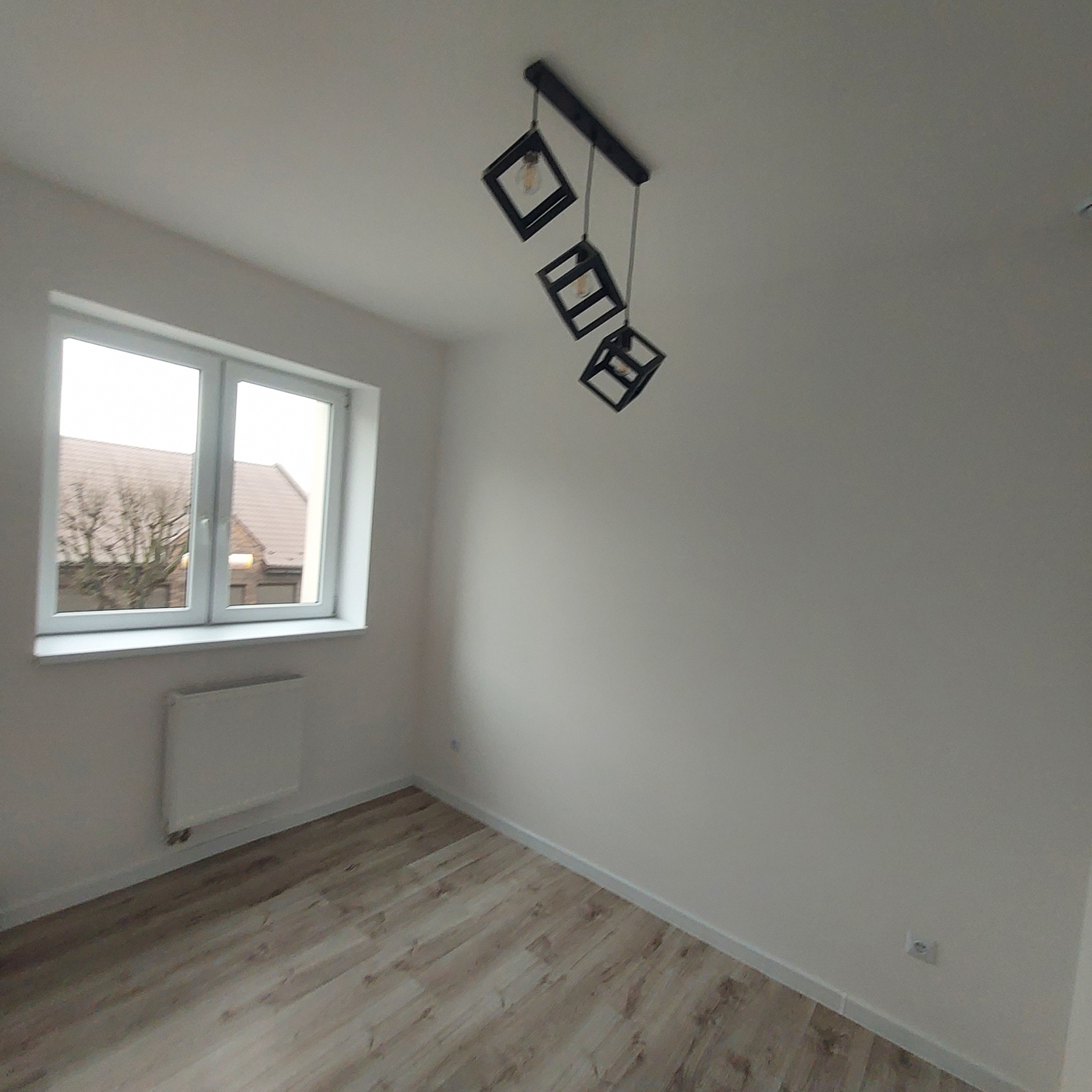 Pusty pokój po remoncie z panelami podłogowymi w odcieniach drewna, pomalowanymi ścianami na biało, grzejnikiem pod oknem i nowoczesną lampą sufitową z trzema kwadratowymi kloszami.