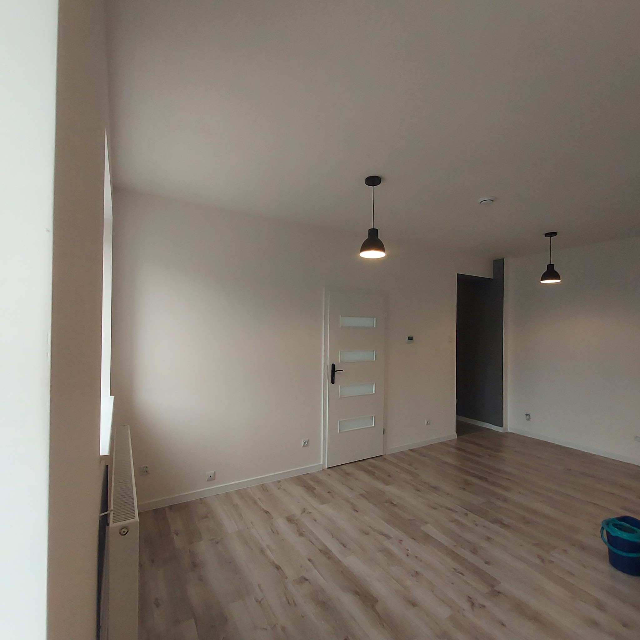 Jasne, puste wnętrze po remoncie z panelami podłogowymi, pomalowanymi ścianami, białą stolarką drzwiową i minimalistycznym oświetleniem, widoczny grzejnik oraz wiadro.