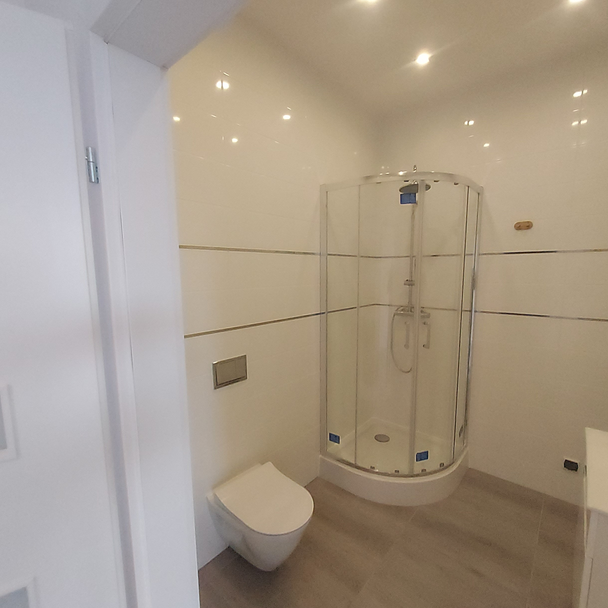 Nowoczesna łazienka w Zelowie po remoncie: białe płytki z poziomymi paskami, kabina prysznicowa narożna ze szklanymi drzwiami, podwieszana toaleta z chromowanym przyciskiem, jasna podłoga imitująca...