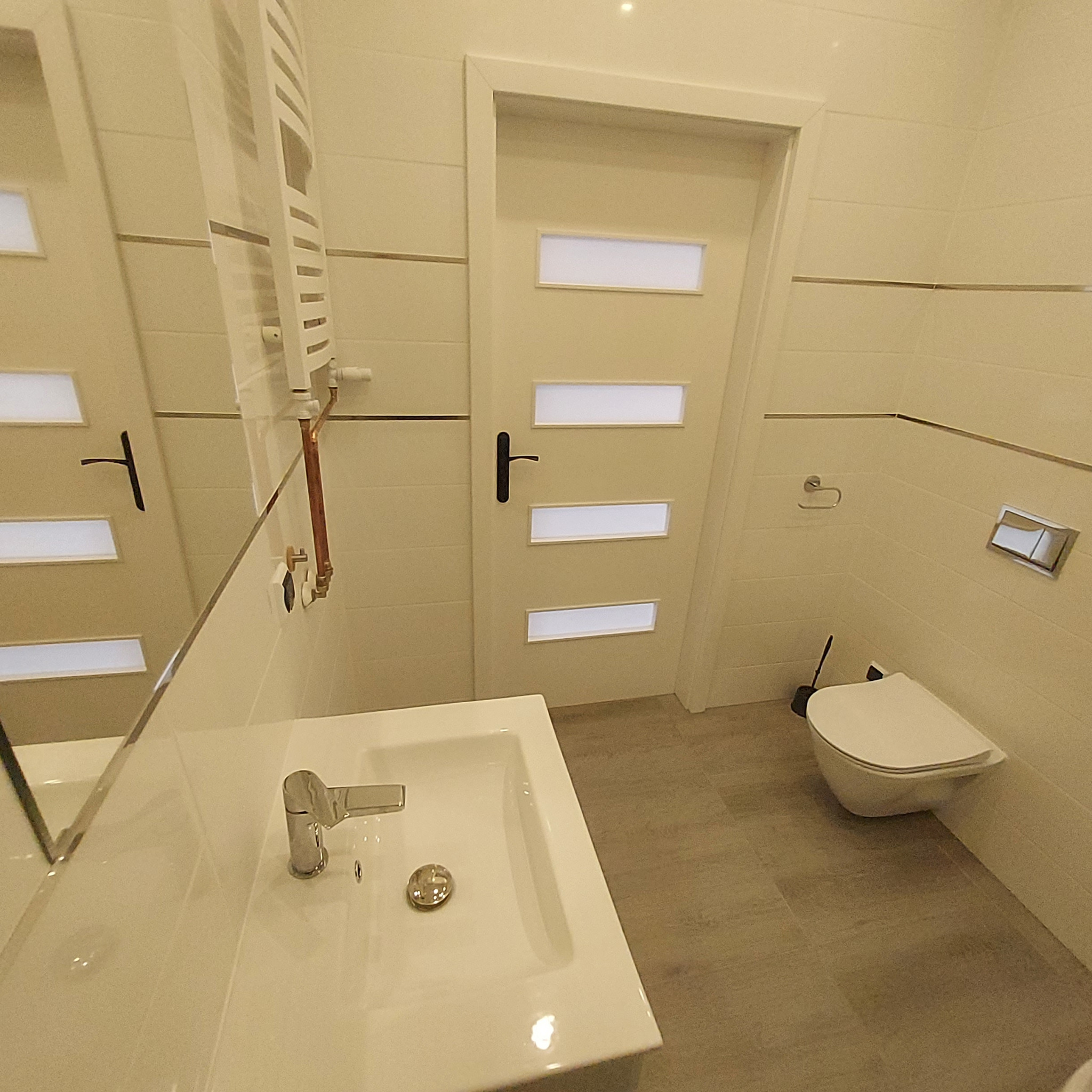 Biała, nowoczesna łazienka z umywalką, toaletą podwieszaną i grzejnikiem drabinkowym, wykończona płytkami w odcieniach beżu z poziomymi, srebrnymi listwami dekoracyjnymi.