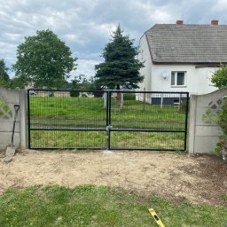 Szymon Brańka - Czarna, metalowa brama panelowa zamontowana między betonowymi słupkami ogrodzenia. Widoczna łopata i poziomica, sugerujące trwający montaż. W tle dom i zieleń.