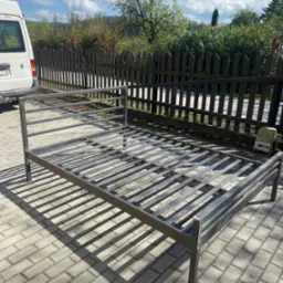 Metalowa konstrukcja łóżka, spawana, na kostce brukowej. W tle płot, zieleń i błękitne niebo. Konstrukcja w surowym stanie, gotowa do dalszej obróbki lub malowania.