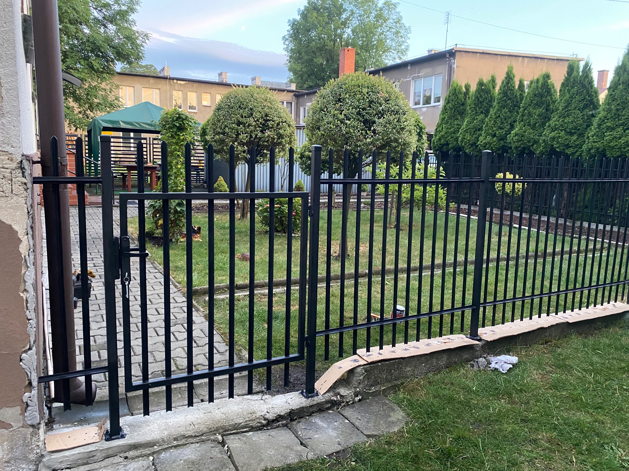 Czarny, metalowy płot panelowy z furtką na tle zielonego trawnika i budynków. Widoczne elementy montażu. Kąt ujęcia od strony posesji.