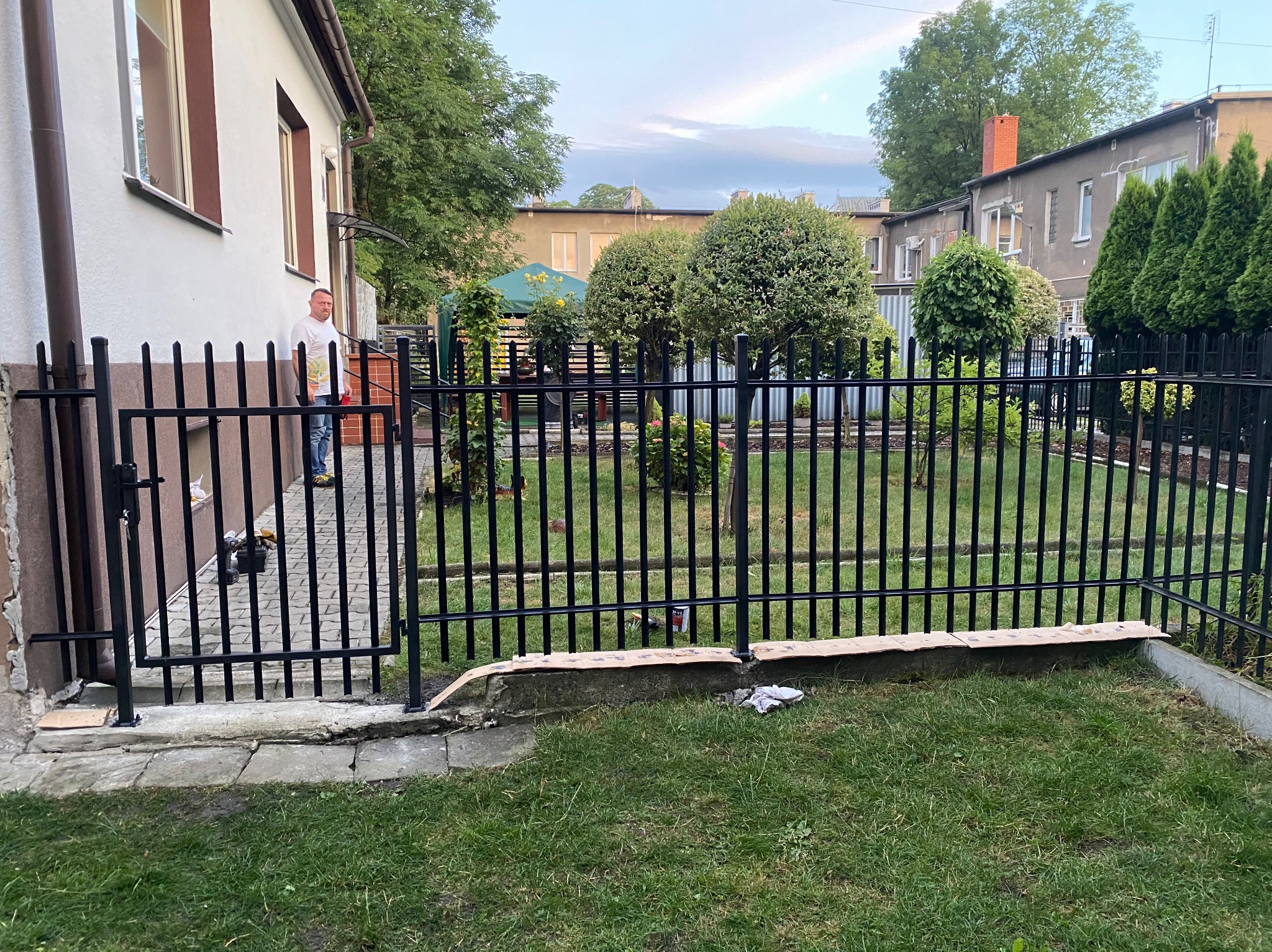 Czarny metalowy płot z furtką, widok na zadbany ogród z kulistymi drzewami. Mężczyzna w tle przy budynku. Elementy montażu widoczne przy podstawie ogrodzenia.