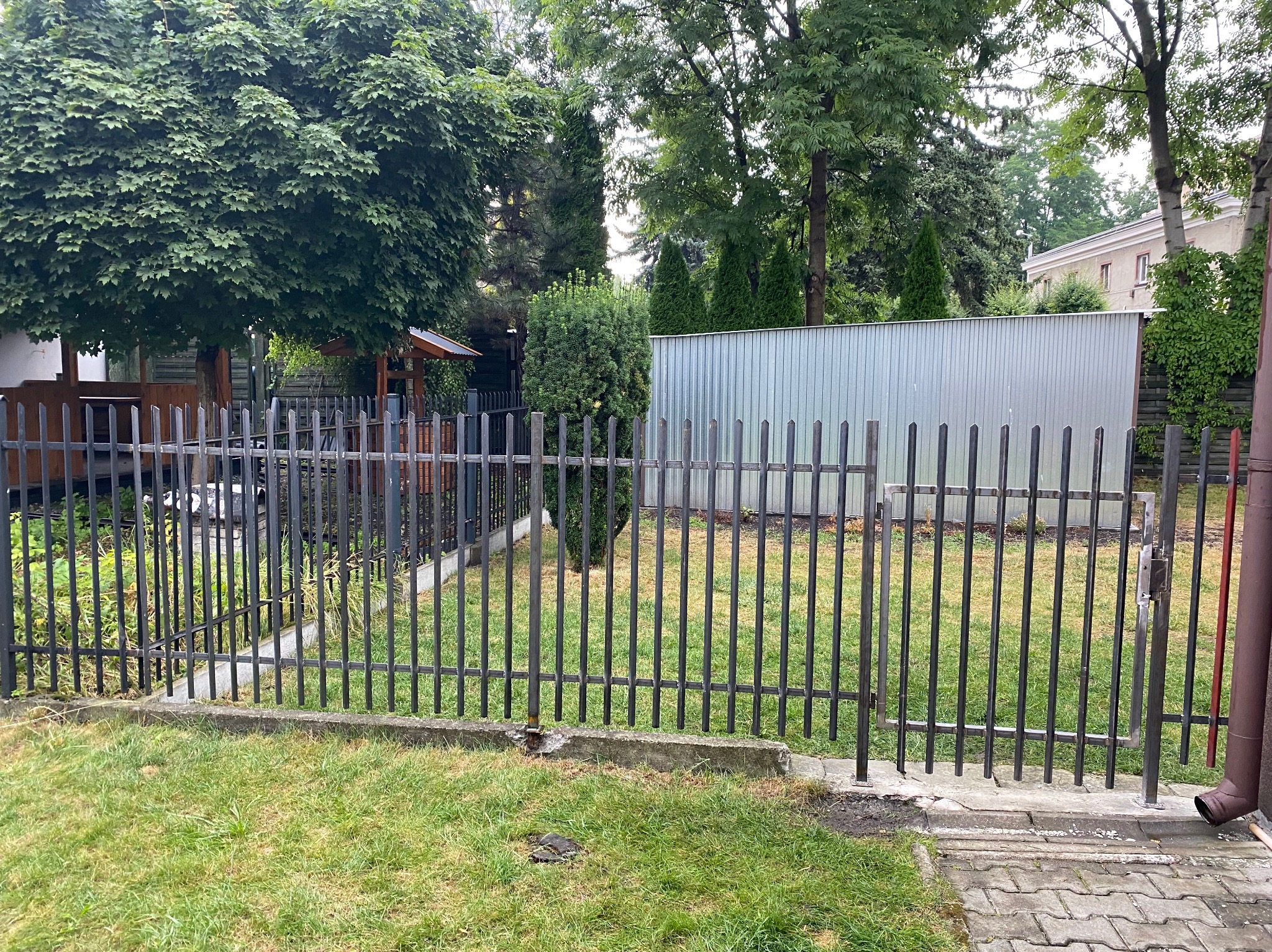 Ciemna, metalowa, sztachetowa furtka ogrodowa na tle zielonego trawnika i drzew. Widoczny fragment posesji z budynkiem i metalowym ogrodzeniem w tle.