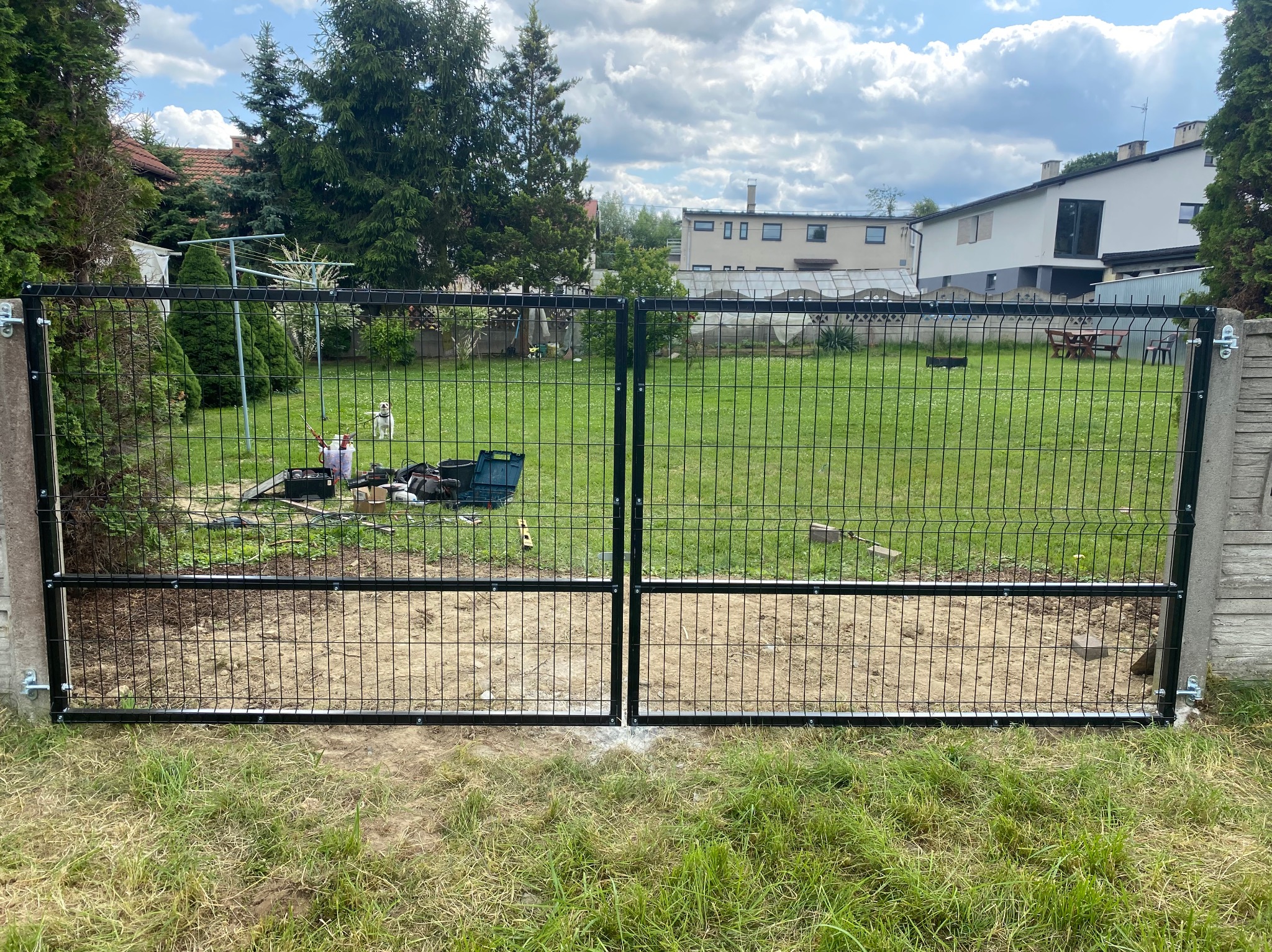 Czarna, dwuskrzydłowa brama panelowa z widocznymi narzędziami montażowymi w tle, osadzona między betonowymi słupkami, z trawnikiem i zabudową w tle.