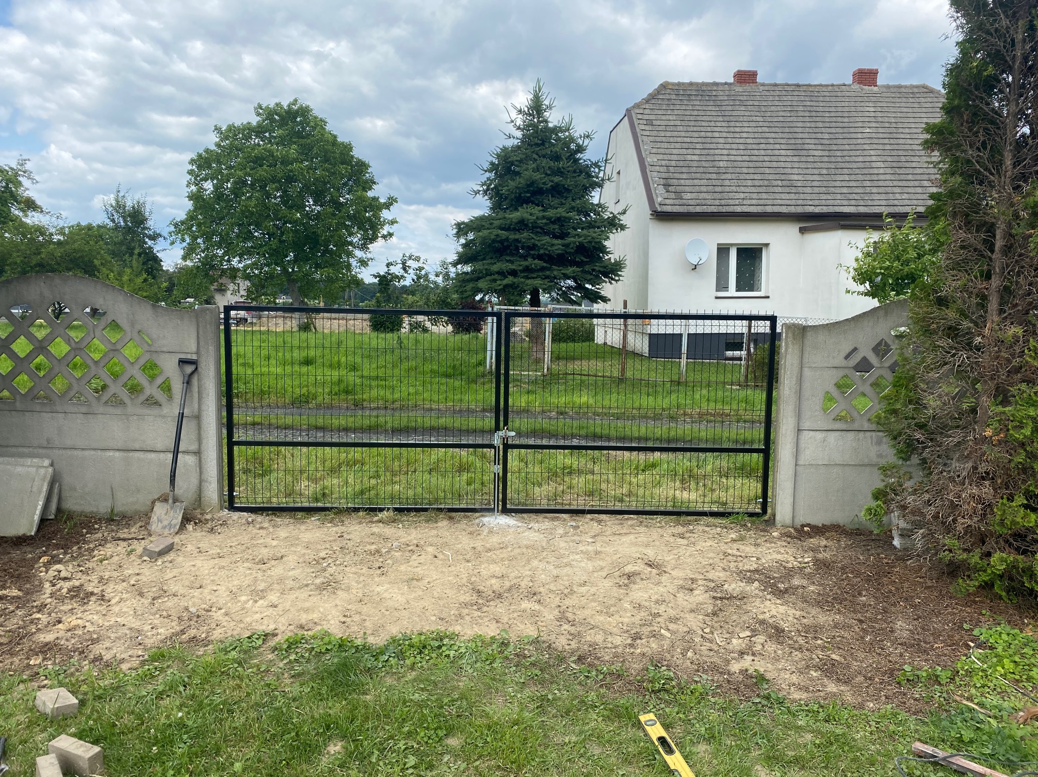 Czarna, metalowa brama panelowa zamontowana między betonowymi słupkami ogrodzenia. Widoczna łopata i poziomica, sugerujące trwający montaż. W tle dom i zieleń.
