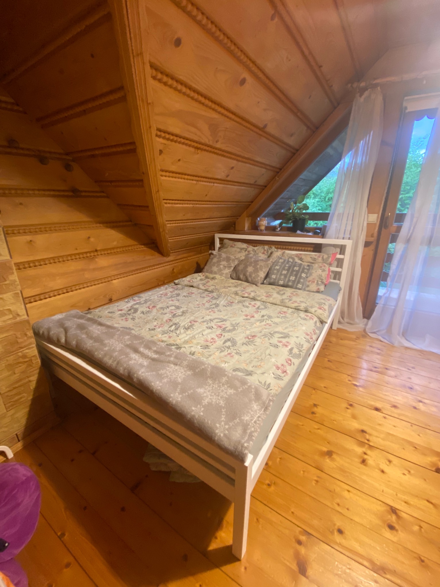 Drewniana sypialnia na poddaszu z białym łóżkiem, kwiatowym wzorem pościeli i oknem z widokiem na zieleń. Charakterystyczne wykończenie ścian i sufitu z frezowanego drewna.