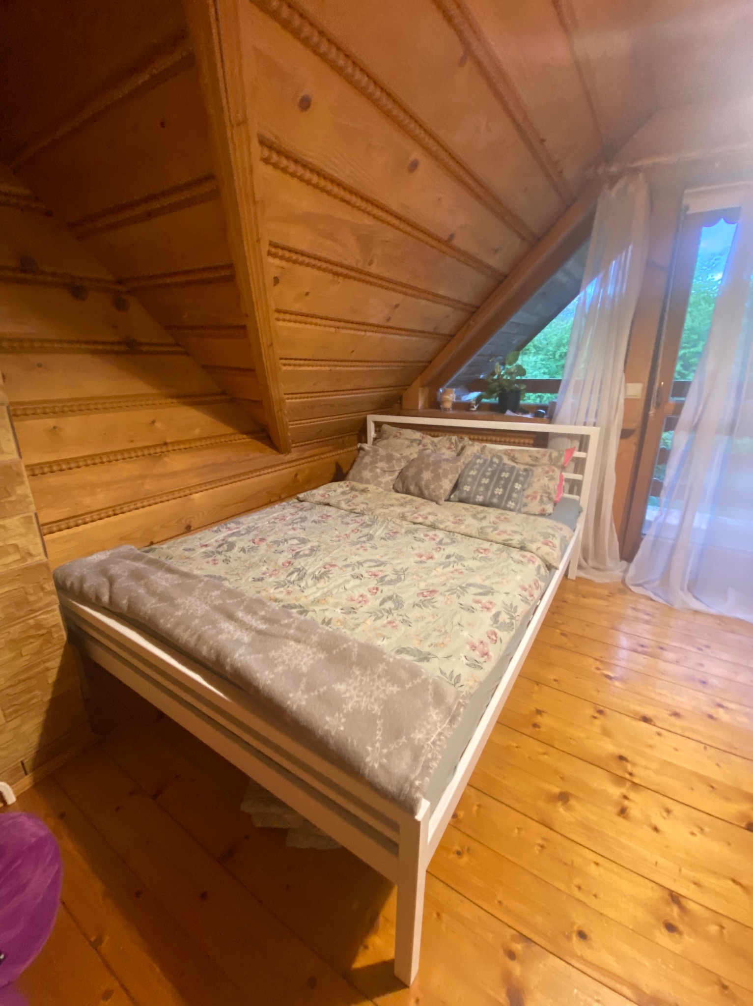 Drewniany pokój na poddaszu z białym łóżkiem i oknem z widokiem na zieleń. Ściany i podłoga wykończone boazerią, co nadaje wnętrzu rustykalny charakter.