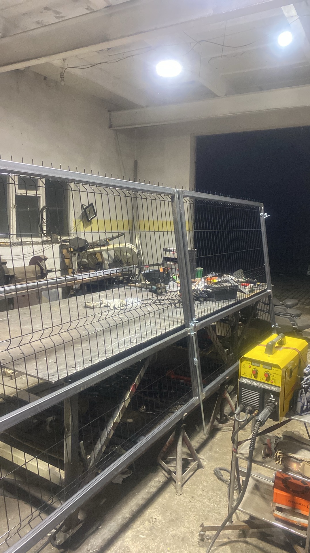 Warsztatowa sceneria: spawarka obok fragmentu ogrodzenia panelowego na metalowym stole. W tle okno i narzędzia, oświetlenie sufitowe.