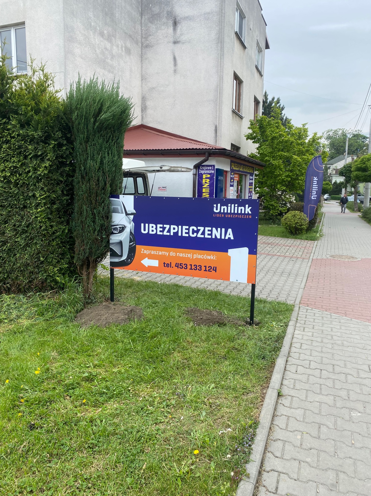 Billboard Unilink Ubezpieczenia z numerem telefonu, częściowo zasłonięty przez krzew. W tle budynek i chodnik z kostki brukowej.
