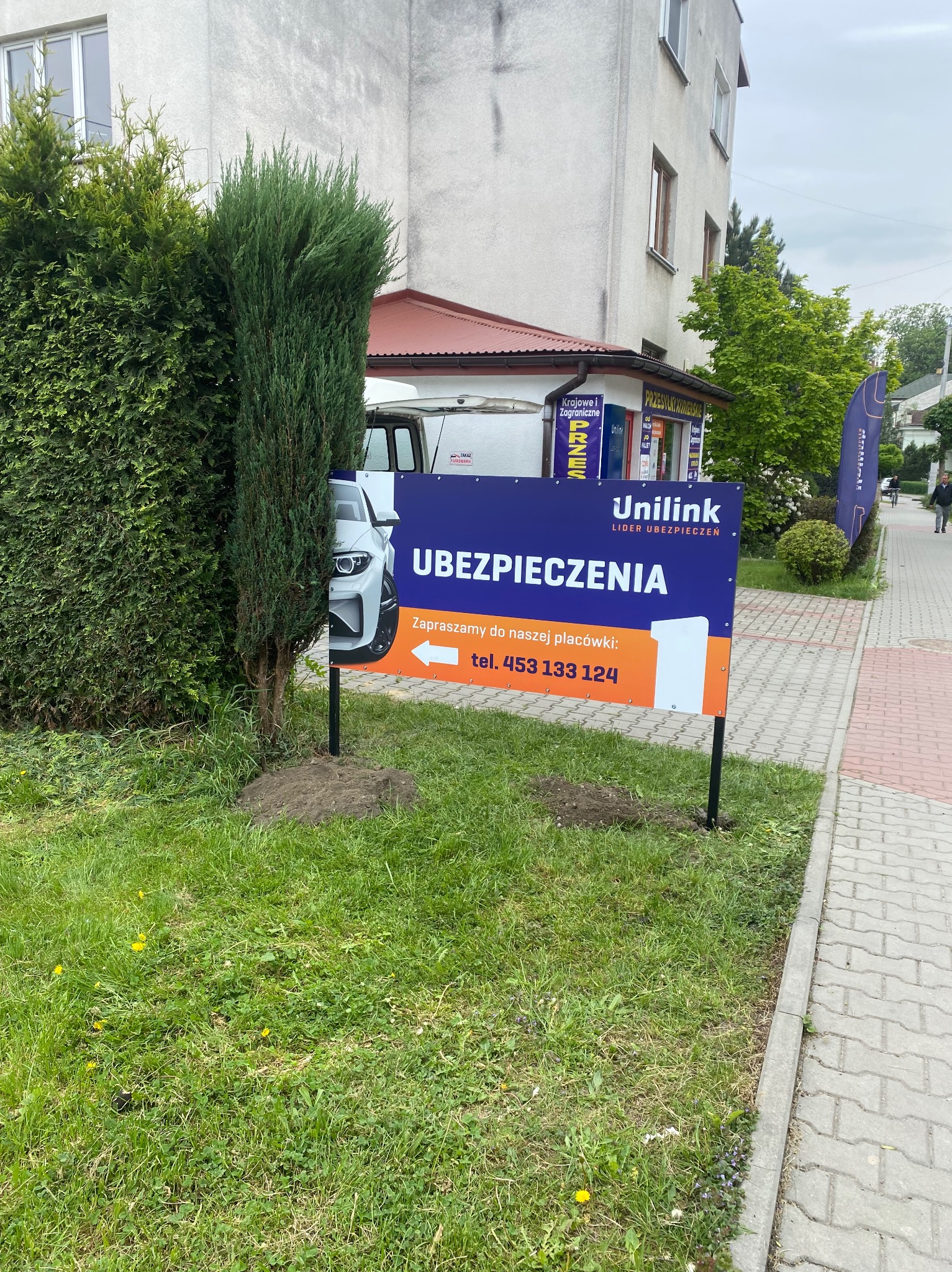 Zewnętrzny baner reklamowy Unilink Ubezpieczenia na trawniku obok chodnika, z częściowo widocznym samochodem i budynkiem w tle. Reklama agencji ubezpieczeniowej.