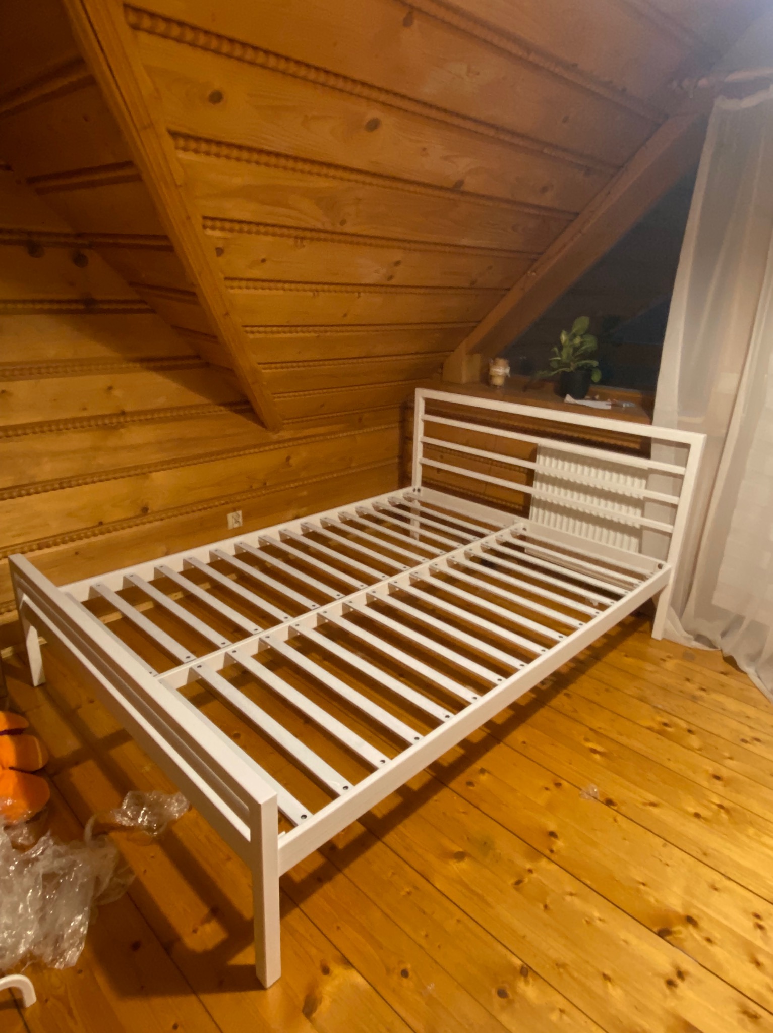 Biała, drewniana rama łóżka w pokoju z boazerią, Wadowice. Widok na stelaż łóżka na tle drewnianej ściany i podłogi. Minimalistyczny design ramy.