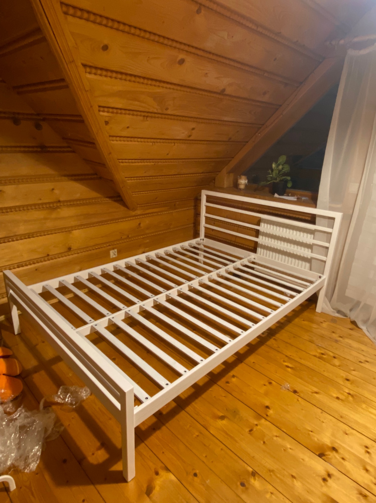 Biała, drewniana rama łóżka bez materaca w pokoju z drewnianą boazerią. Widoczne elementy montażowe i okno z firanką.