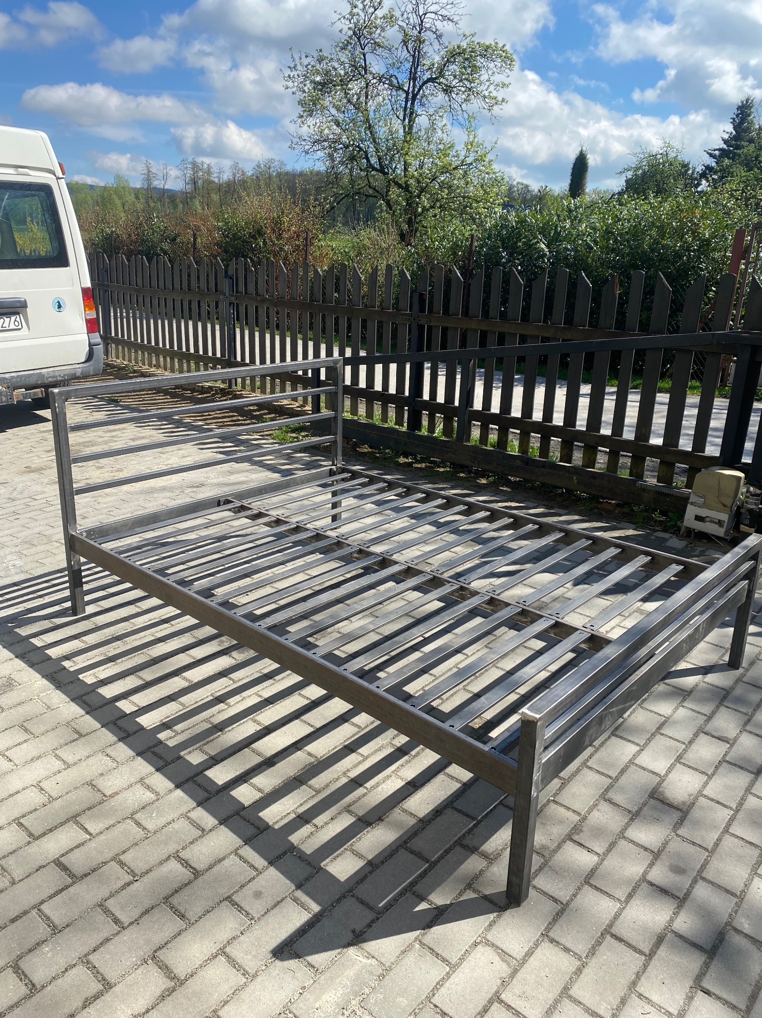 Metalowa konstrukcja łóżka, spawana, na kostce brukowej. W tle płot, zieleń i błękitne niebo. Konstrukcja w surowym stanie, gotowa do dalszej obróbki lub malowania.