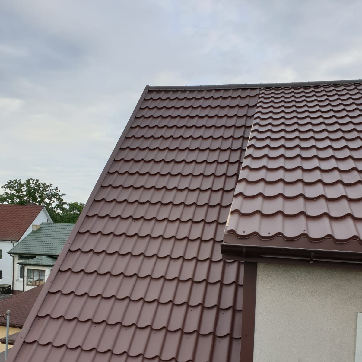 Nowy, brązowy dach z blachodachówki modułowej, z zamontowaną rynną, na tle pochmurnego nieba i zabudowań sąsiednich.