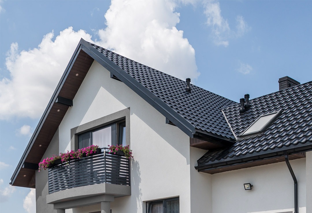 Nowoczesny dom jednorodzinny z ciemną dachówką ceramiczną, balkonem z kwiatami i oknami dachowymi, na tle błękitnego nieba z chmurami.