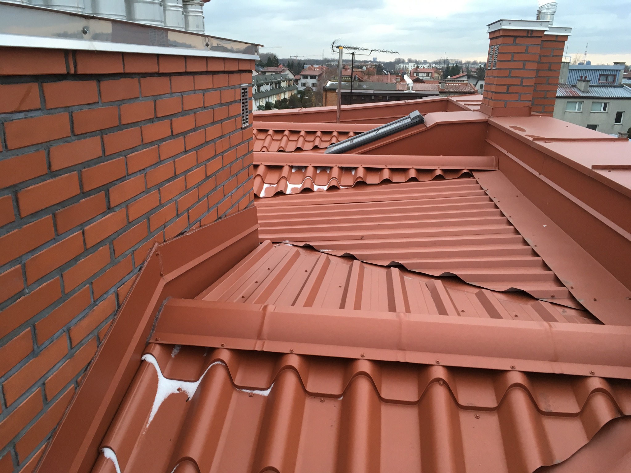 Nowy, pomarańczowo-czerwony dach z blachy trapezowej i dachówki ceramicznej, z widocznym kominem z cegły i fragmentami śniegu w załamaniach dachówki, na tle zabudowy miejskiej pod pochmurnym niebem.