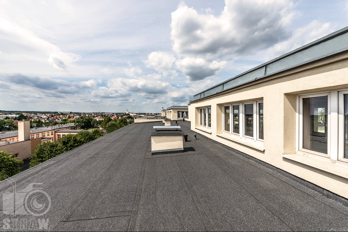 Widok z góry na płaski dach pokryty papą termozgrzewalną, z widocznymi kominkami wentylacyjnymi i budynkiem z oknami w tle, na tle pochmurnego nieba i panoramy miasta.