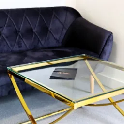Stylowy, granatowy, welurowy narożnik z pikowaniami i guziczkami, obok stolik kawowy ze złotym, geometrycznym stelażem i szklanym blatem, na kt&oacute;rym leżą czarne wizyt&oacute;wki.