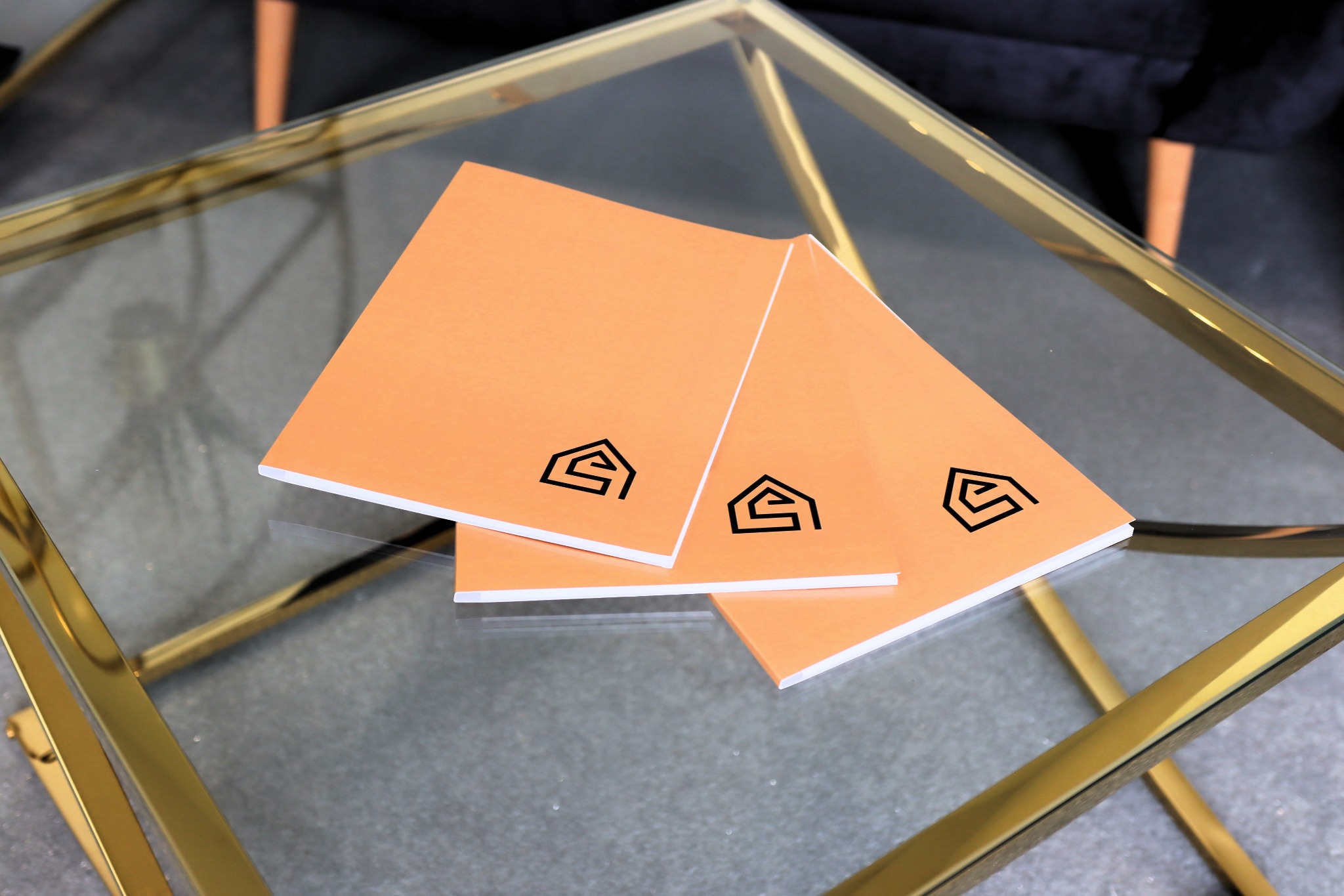 Trzy pomarańczowe kartki z czarnym logo w kształcie domu leżące na szklanym stoliku kawowym ze złotą ramą.