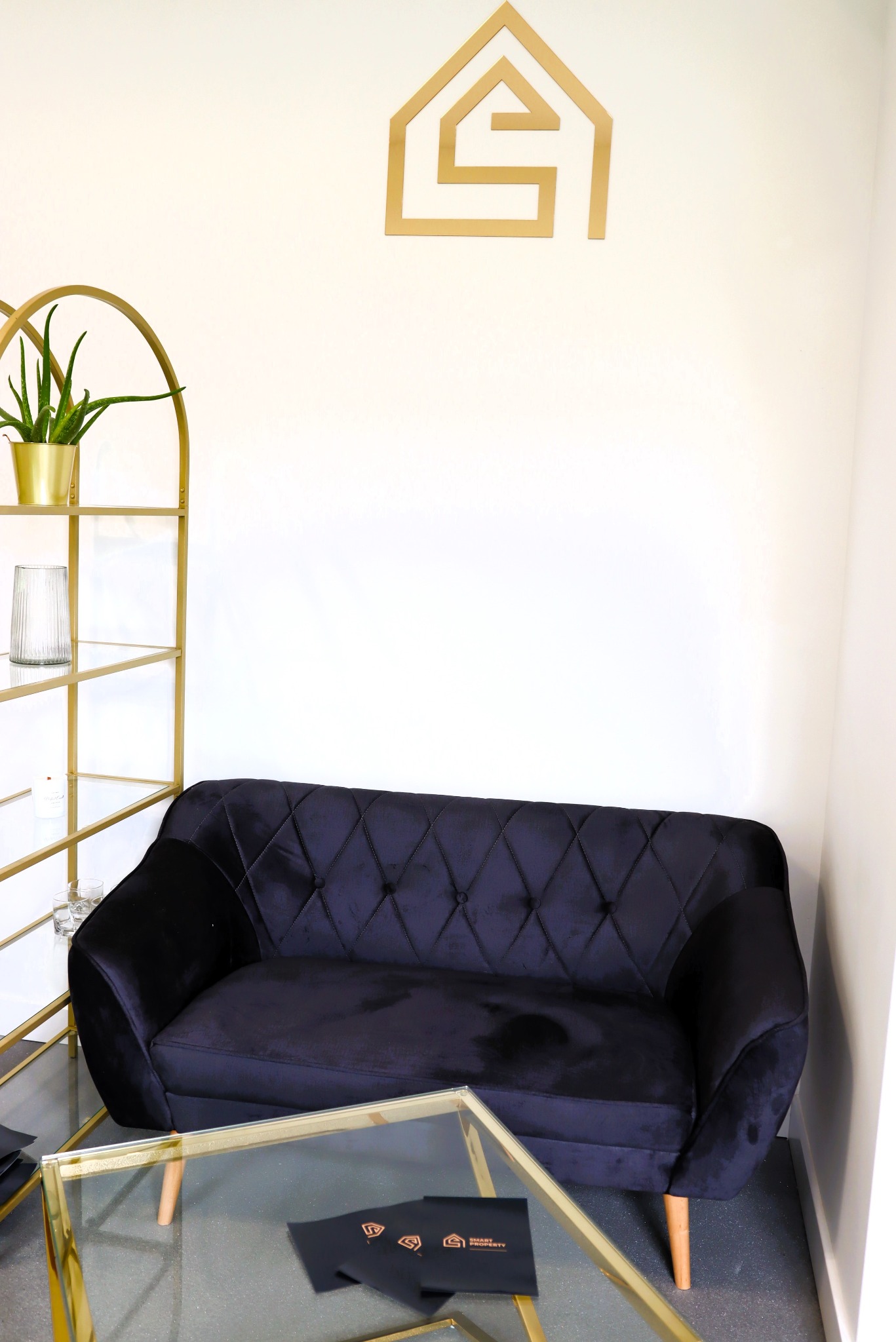 Ciemnoniebieska, pikowana sofa z drewnianymi nóżkami, szklany stolik kawowy ze złotą ramą, złoty regał z rośliną w doniczce i szklanym wazonem, logo firmy na ścianie.