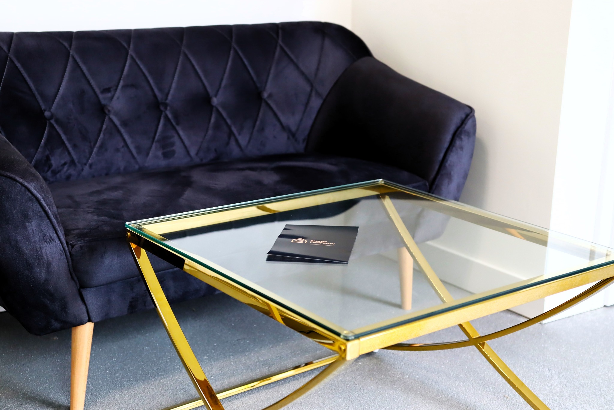 Stylowy, granatowy, welurowy narożnik z pikowaniami i guziczkami, obok stolik kawowy ze złotym, geometrycznym stelażem i szklanym blatem, na którym leżą czarne wizytówki.