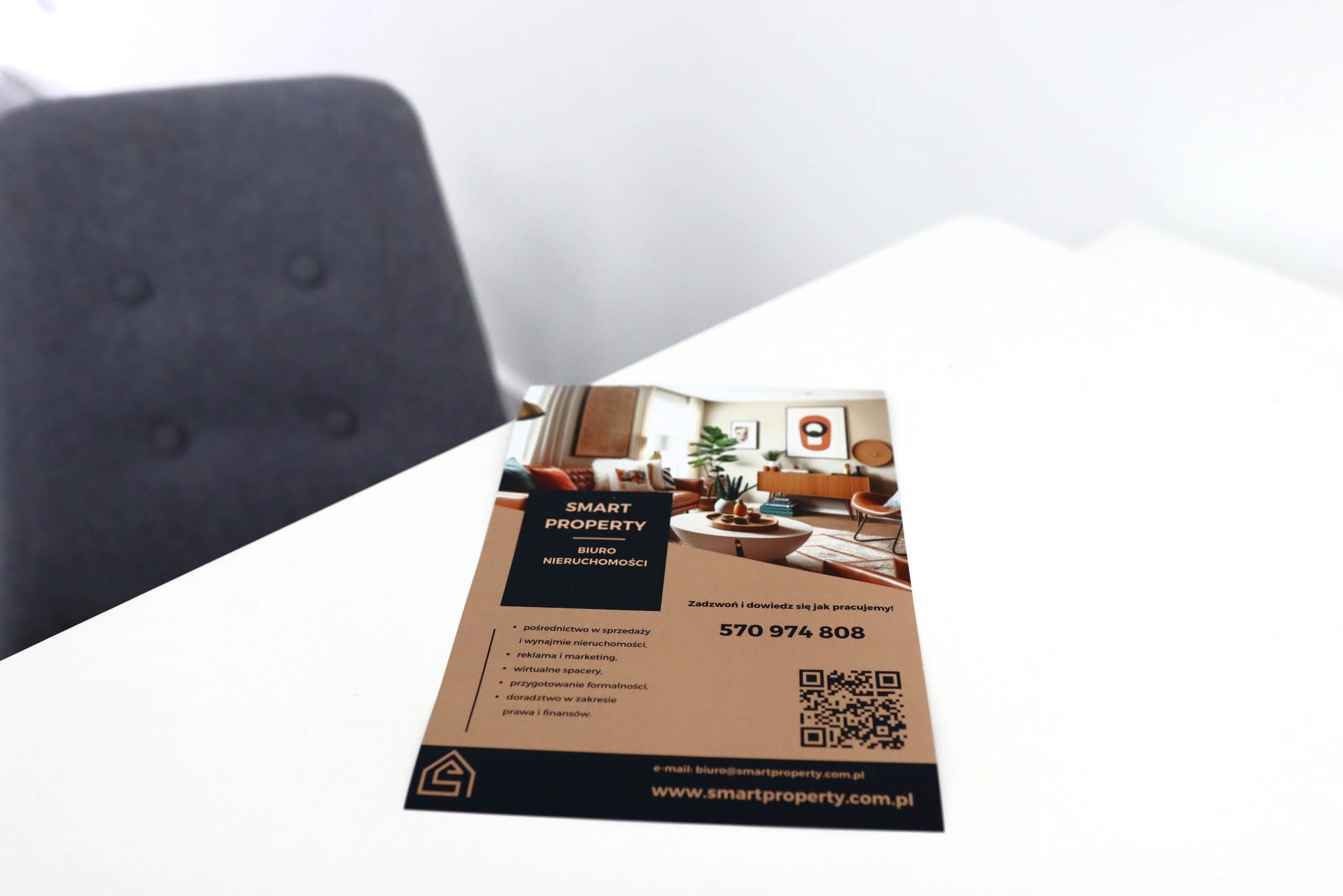 Ulotka biura nieruchomości Smart Property leżąca na białym stole, z widocznym fragmentem szarego krzesła w tle, prezentująca ofertę pośrednictwa w sprzedaży i wynajmie, reklamę, wirtualne spacery...
