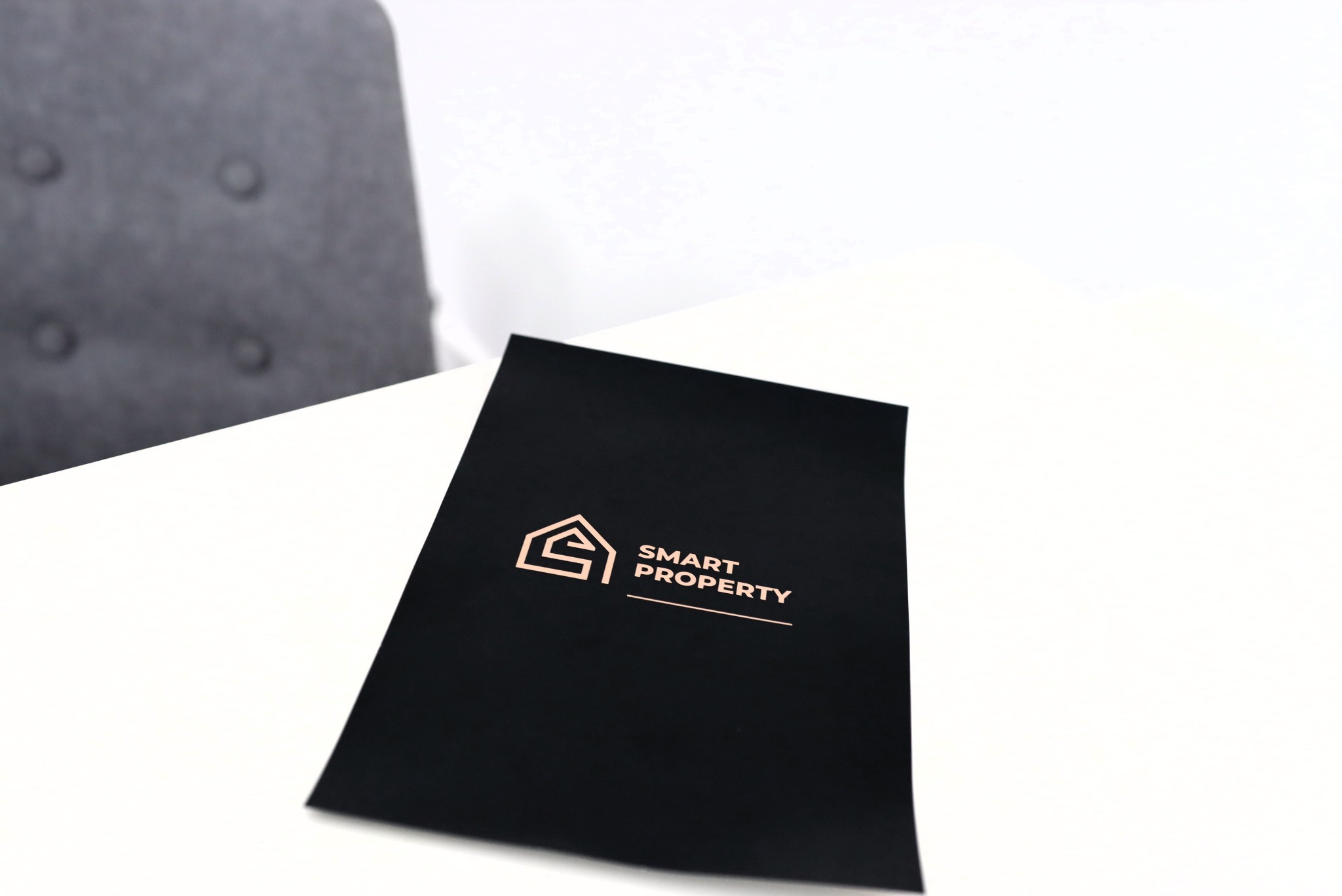 Czarna teczka z logo Smart Property w kolorze różowego złota, leżąca na białym stole, z fragmentem szarego krzesła w tle.