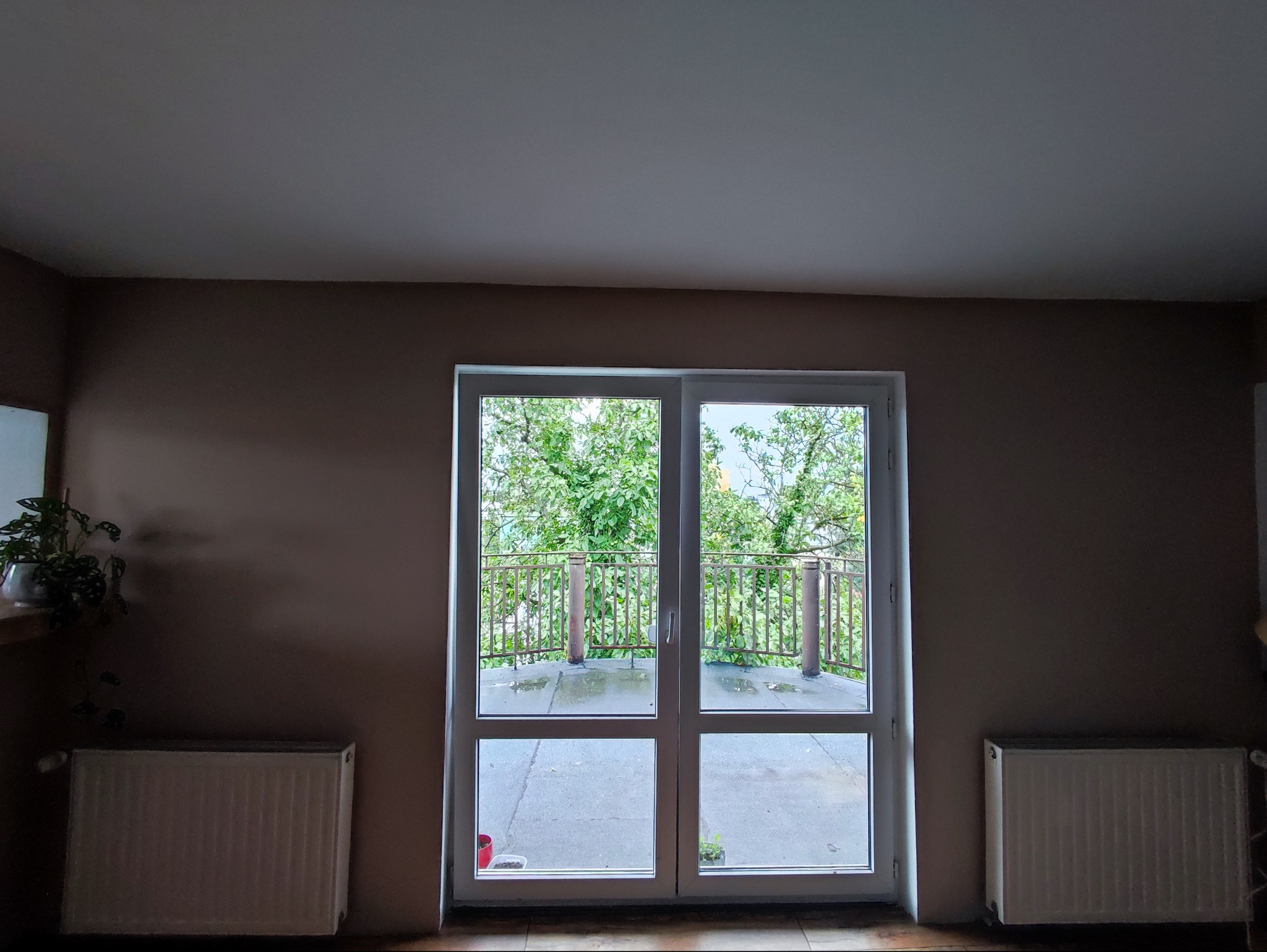 Pokój po malowaniu ścian na beżowo, z widokiem na balkon z zielenią przez duże okno balkonowe.