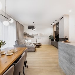 ZWOLMAR MEBLE NA WYMIAR - Jasne wnętrze z drewnianym stołem jadalnym, szarą sofą narożną i wyspą kuchenną z blatem imitującym beton, oświetlone nowoczesnymi lampami.
