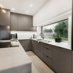 ZWOLMAR MEBLE NA WYMIAR - Nowoczesna kuchnia z szarymi szafkami, białym blatem i dużym oknem z widokiem na ogród, oświetlona lampą sufitową z kilkoma żarówkami w stylu retro.