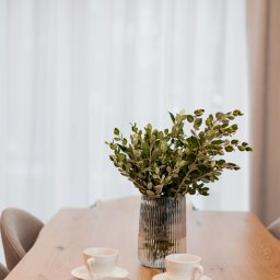 ZWOLMAR MEBLE NA WYMIAR - Drewniany stół jadalny z blatem z widocznymi słojami, nakryty białymi filiżankami i talerzami, dekorowany szklanym wazonem z zielonymi gałązkami, oświetlony lampą sufitową na tle białej zasłony.