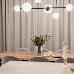 ZWOLMAR MEBLE NA WYMIAR - Jasny pokój jadalny z drewnianym stołem, szarymi krzesłami, nowoczesną lampą wiszącą z białymi kulami i wazonem z zielonymi gałązkami na stole, nakryty do posiłku.