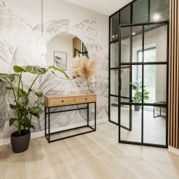 ZWOLMAR MEBLE NA WYMIAR - Stylowy przedpokój w Bydgoszczy z konsolą z dwiema szufladami, okrągłym lustrem i dekoracyjną ścianą z motywem roślinnym. W tle widoczne drzwi w stylu loftowym z czarnymi szprosami i drewniane lamele.