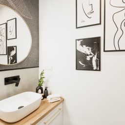 ZWOLMAR MEBLE NA WYMIAR - Nowoczesna łazienka z owalnym lustrem w złotej ramie, umywalką nablatową, czarną armaturą, drewnianym blatem i minimalistycznymi grafikami na ścianie.