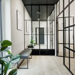 Nowoczesny przedpok&oacute;j z czarną, przeszkloną ścianką działową, szarą ławką i rośliną doniczkową przy oknie.