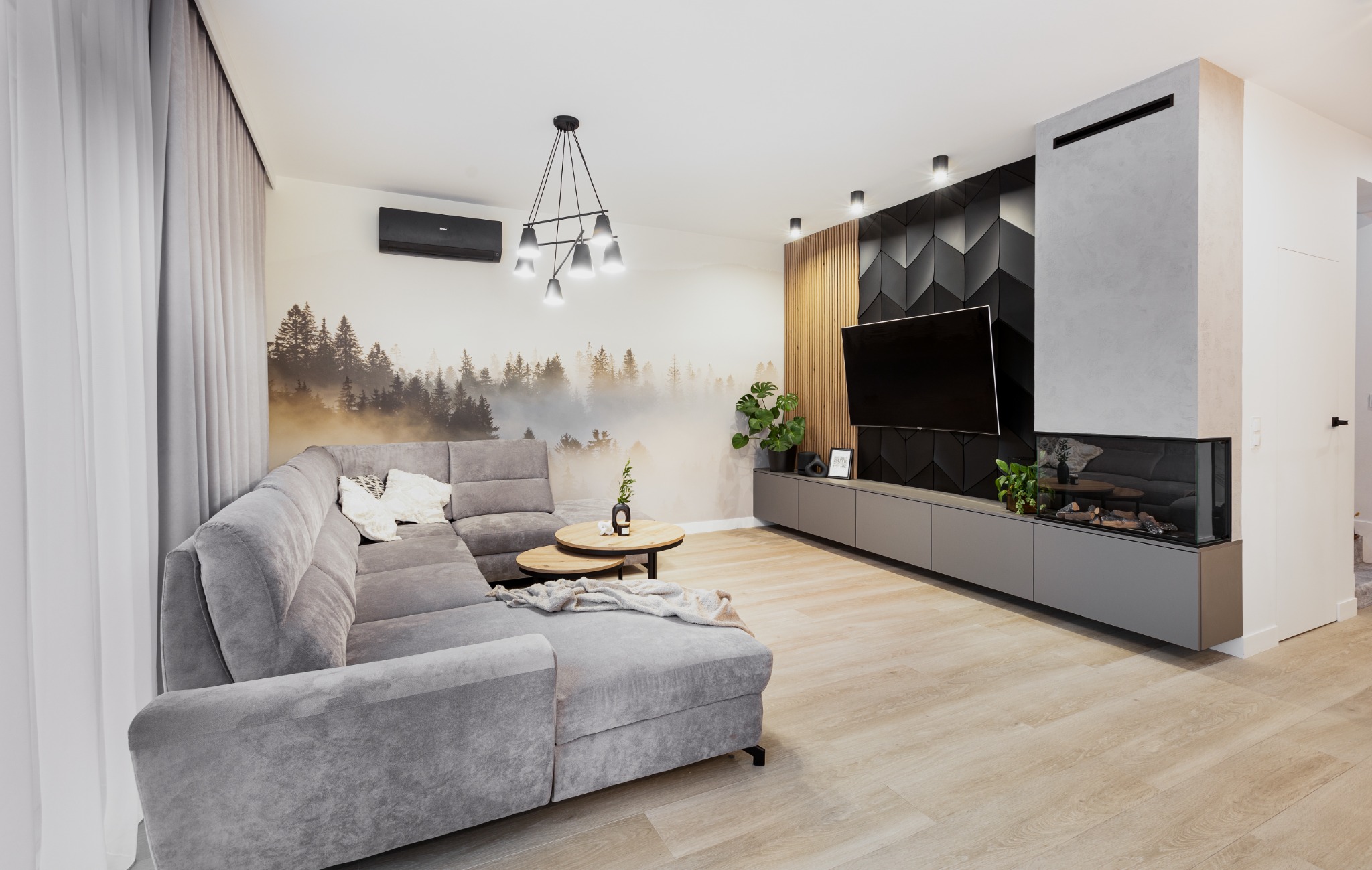 Nowoczesny salon z szarą narożną sofą, okrągłymi stolikami kawowymi i ścianą TV z kominkiem elektrycznym, oświetlony wiszącą lampą z żarówkami Edison.