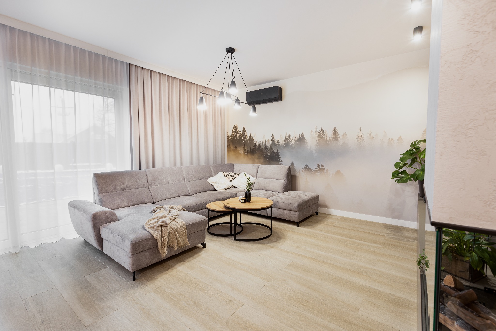 Jasnoszary narożnik z pufem i pledem w nowoczesnym salonie z drewnianą podłogą, okrągłymi stolikami kawowymi i fototapetą z motywem lasu we mgle.