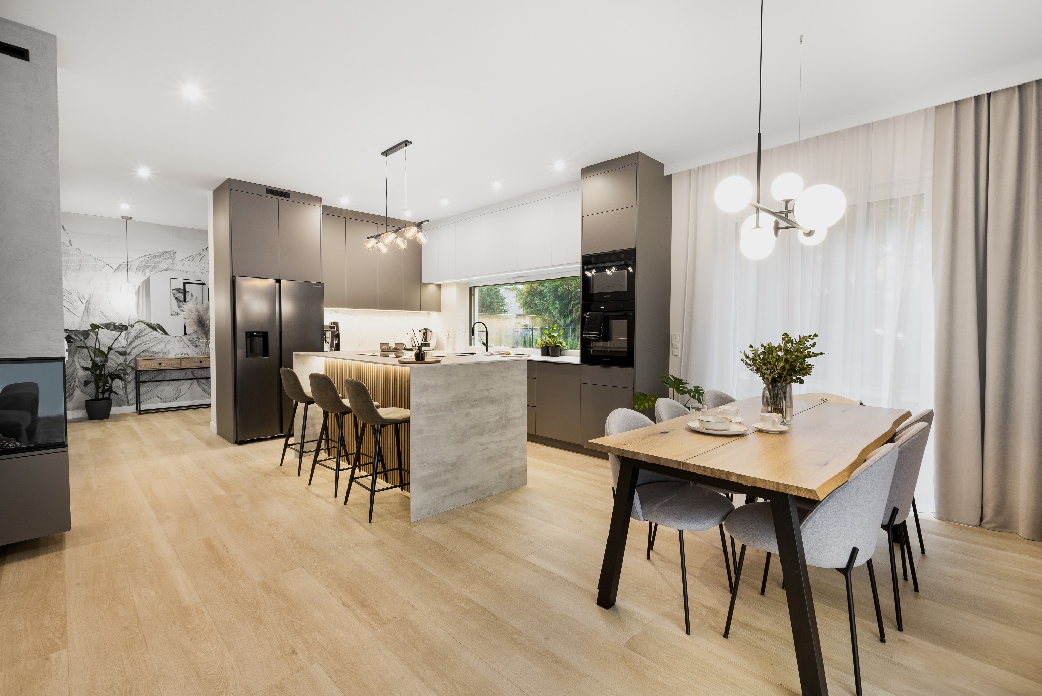 Nowoczesna kuchnia z wyspą, szare szafki, drewniane blaty i krzesła barowe, stół z szarymi krzesłami, jasna podłoga i duże okno z widokiem na zieleń.