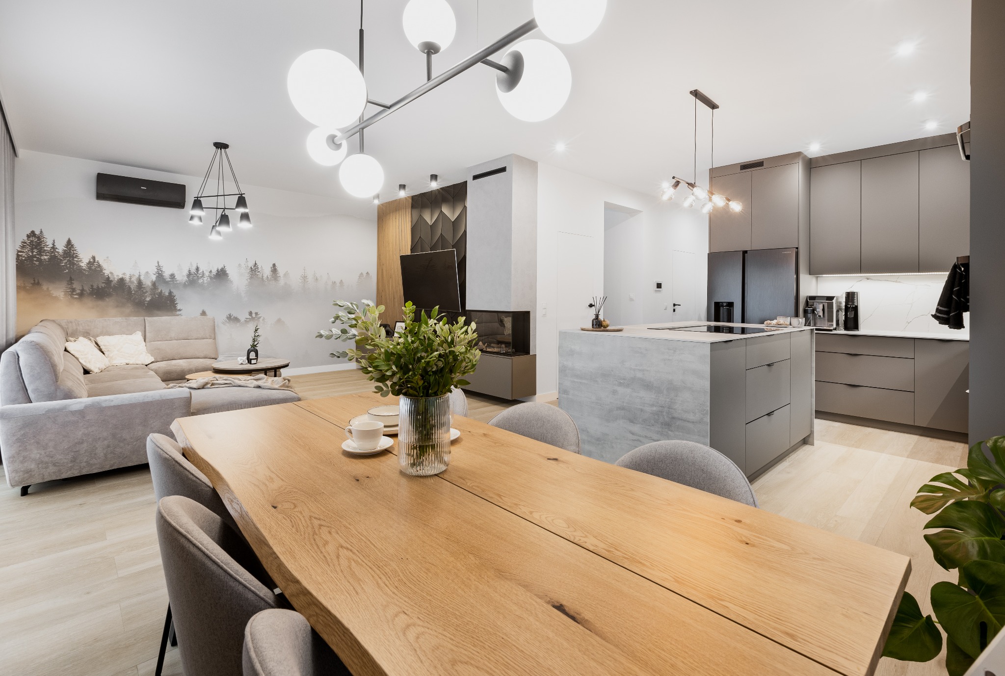 Jasne, przestronne wnętrze z drewnianym stołem na pierwszym planie, szarą narożną sofą z poduszkami i fototapetą z motywem lasu w tle oraz nowoczesną szarą zabudową kuchenną z wyspą i marmurowym...