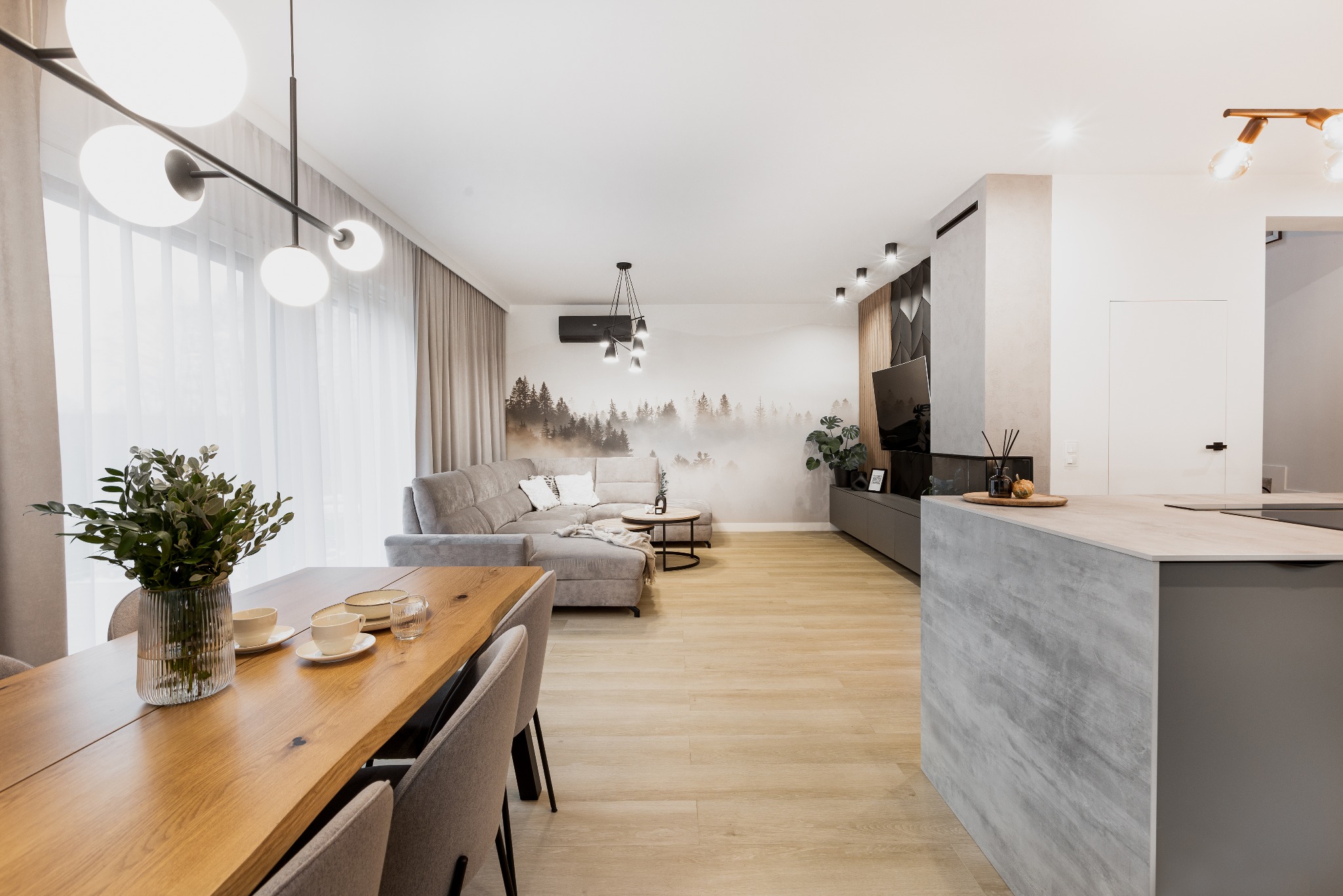 Jasne wnętrze z drewnianym stołem jadalnym, szarą sofą narożną i wyspą kuchenną z blatem imitującym beton, oświetlone nowoczesnymi lampami.