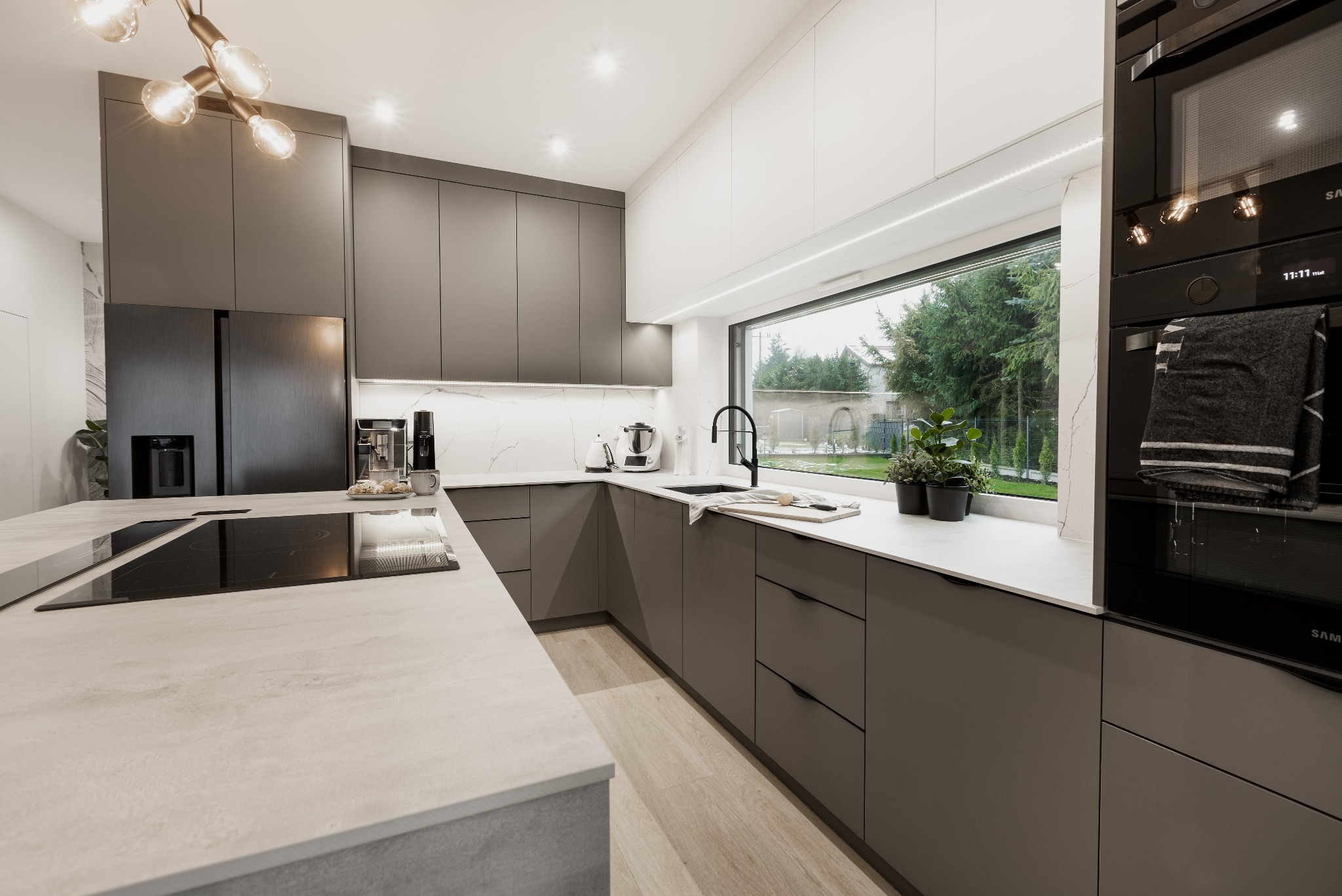 Nowoczesna kuchnia z szarymi szafkami, białym blatem i dużym oknem z widokiem na ogród, oświetlona lampą sufitową z kilkoma żarówkami w stylu retro.
